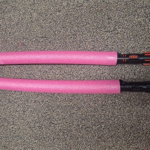 May include: Two pink foam light sabers with black and orange handles. The light sabers are lying on a textured, gray surface. The handles have a detailed design.