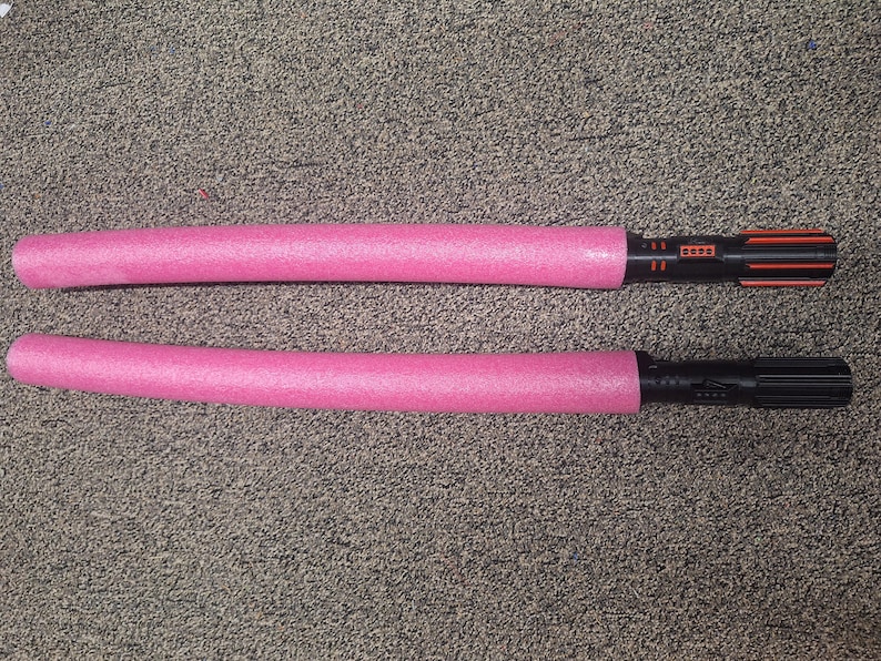 May include: Two pink foam light sabers with black and orange handles. The light sabers are lying on a textured, gray surface. The handles have a detailed design.