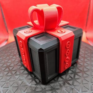 May include: A 3D-printed gift box with a red bow and accents. The box is black with red bands and decorative details. The bow is a prominent feature, adding a festive touch to the design. The box sits on a textured surface.
