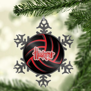 May include: A silver snowflake ornament with a black and red volleyball design. The volleyball has the word "Huskers" in red script. The ornament is hanging from a silver loop and is set against a blurred green background.