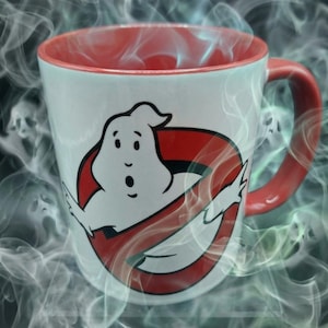 May include: A white ceramic mug with a red interior and handle, featuring a graphic of a white ghost character inside a red circle with a diagonal line across it, surrounded by swirling wisps of gray and green smoke against a dark background.