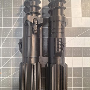 May include: Two black, 3D-printed lightsaber replicas. Each has a textured handle, a detailed midsection with buttons and panels, and a ribbed top. The lightsabers are displayed side-by-side, showcasing their intricate designs.