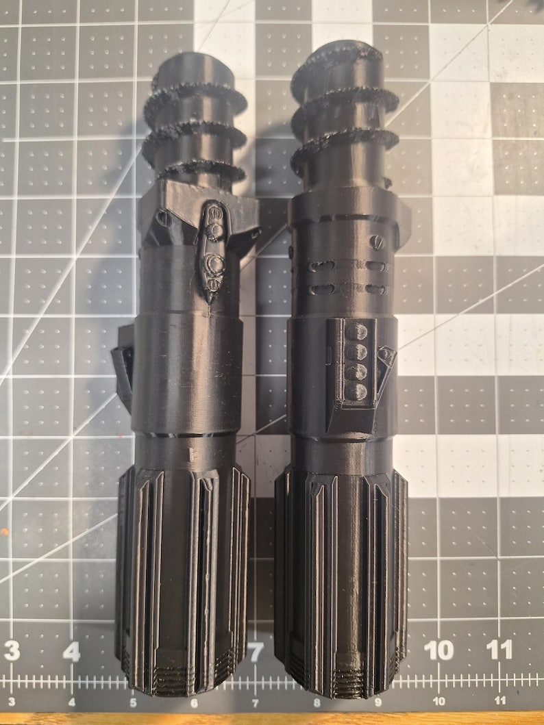 May include: Two black, 3D-printed lightsaber replicas. Each has a textured handle, a detailed midsection with buttons and panels, and a ribbed top. The lightsabers are displayed side-by-side, showcasing their intricate designs.