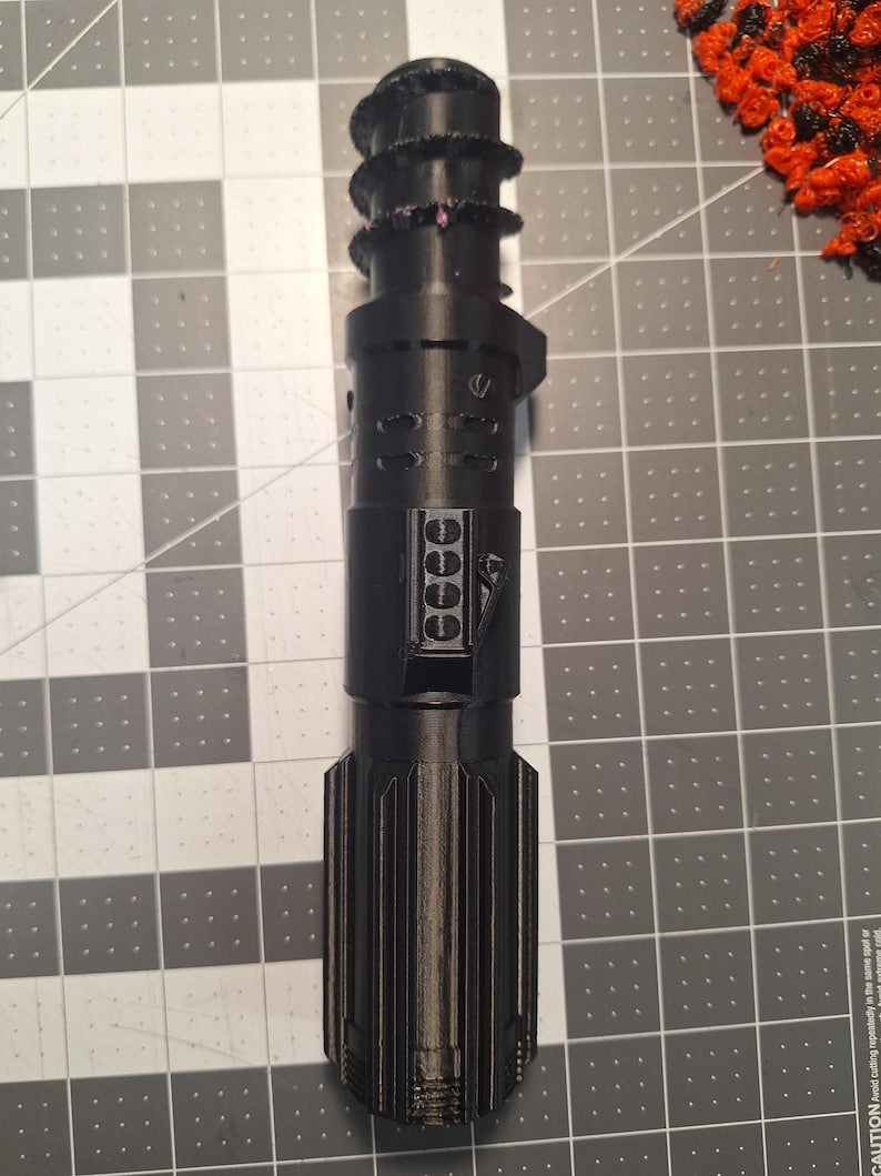 May include: A black, futuristic-looking device with a cylindrical body and textured grip. The device has multiple sections with detailed designs, including a series of small, circular features and a ribbed lower portion. The overall aesthetic suggests a sci-fi or fantasy theme.