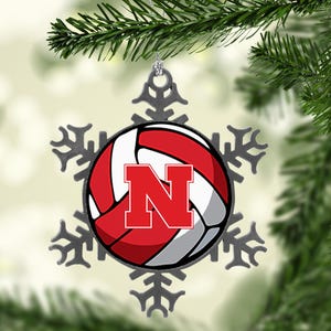 May include: A decorative Christmas ornament featuring a volleyball design with a red "N" logo. The ornament is shaped like a snowflake and is hanging from a silver string. The background is a blurred image of a Christmas tree.