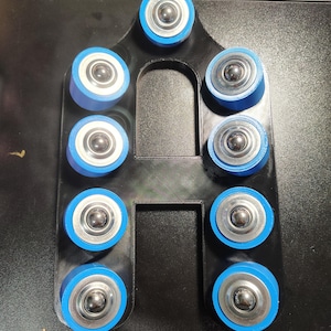May include: A black, arch-shaped frame featuring eight circular, blue-rimmed ball bearing rollers arranged in pairs along the sides and one at the top. The structure has a smooth, matte finish with a central cutout and metallic silver centers.