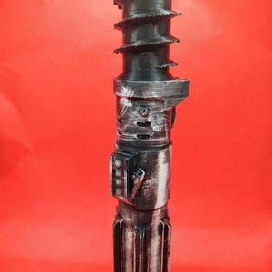May include: A black and silver lightsaber prop with a textured design. The top features a spiral design, and the body has a detailed, metallic appearance. The prop is set against a red background.