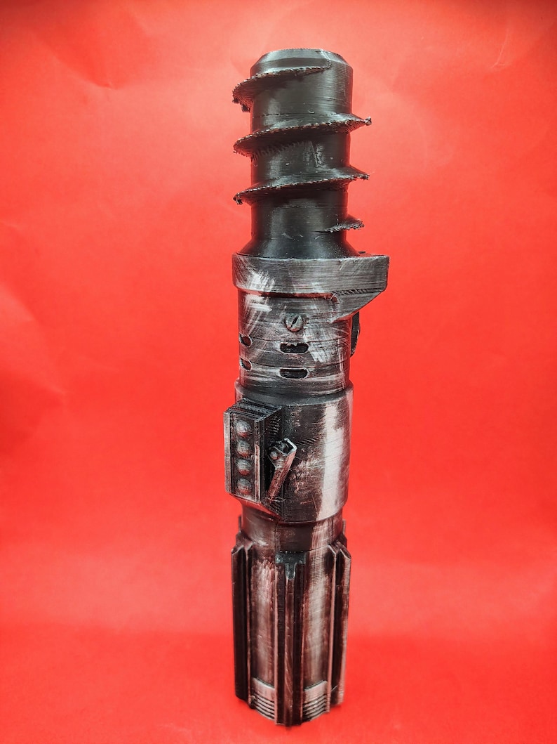 May include: A black and silver lightsaber prop with a textured design. The top features a spiral design, and the body has a detailed, metallic appearance. The prop is set against a red background.