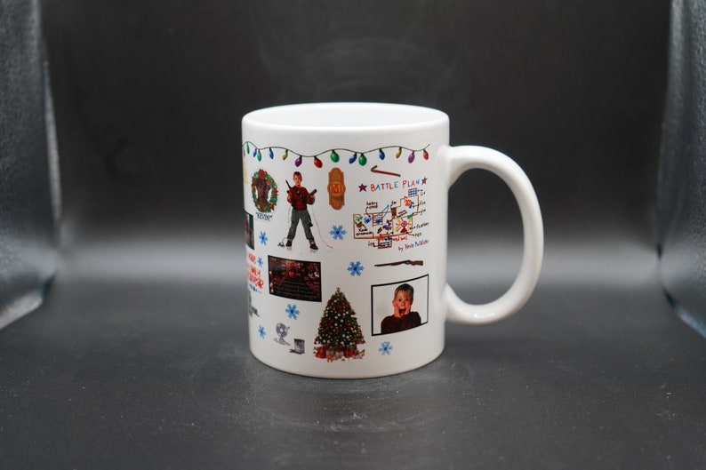 May include: White ceramic mug with a colorful design featuring images and text from the movie Home Alone. The design includes a Christmas tree, a wreath, a string of lights, a map, and a photo of Kevin McCallister.
