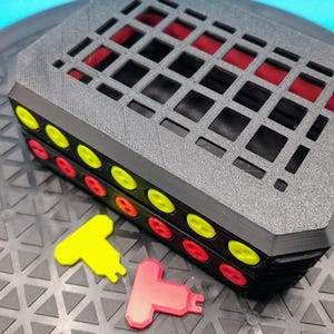 May include: A black, rectangular, 3D-printed object with a grid-like top and red interior. Two rows of yellow and red circular accents are visible on the sides. Two T-shaped pieces, one yellow and one red, are in front.