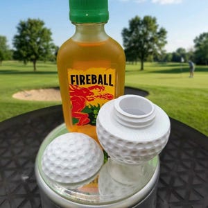 May include: A bottle of Fireball cinnamon whiskey with a green cap, a white golf ball, and a white golf ball-shaped bottle stopper. The items are on a reflective surface with a golf course in the background.