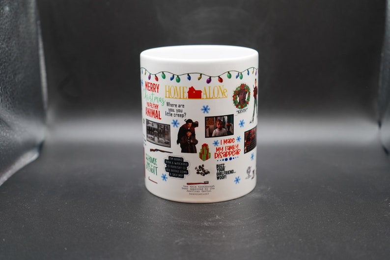 May include: White ceramic mug with a colorful design featuring the movie Home Alone. The mug has a festive Christmas theme with lights, snowflakes, and a wreath. It includes quotes from the movie, such as "Merry Christmas ya filthy animal" and "I made my family disappear."