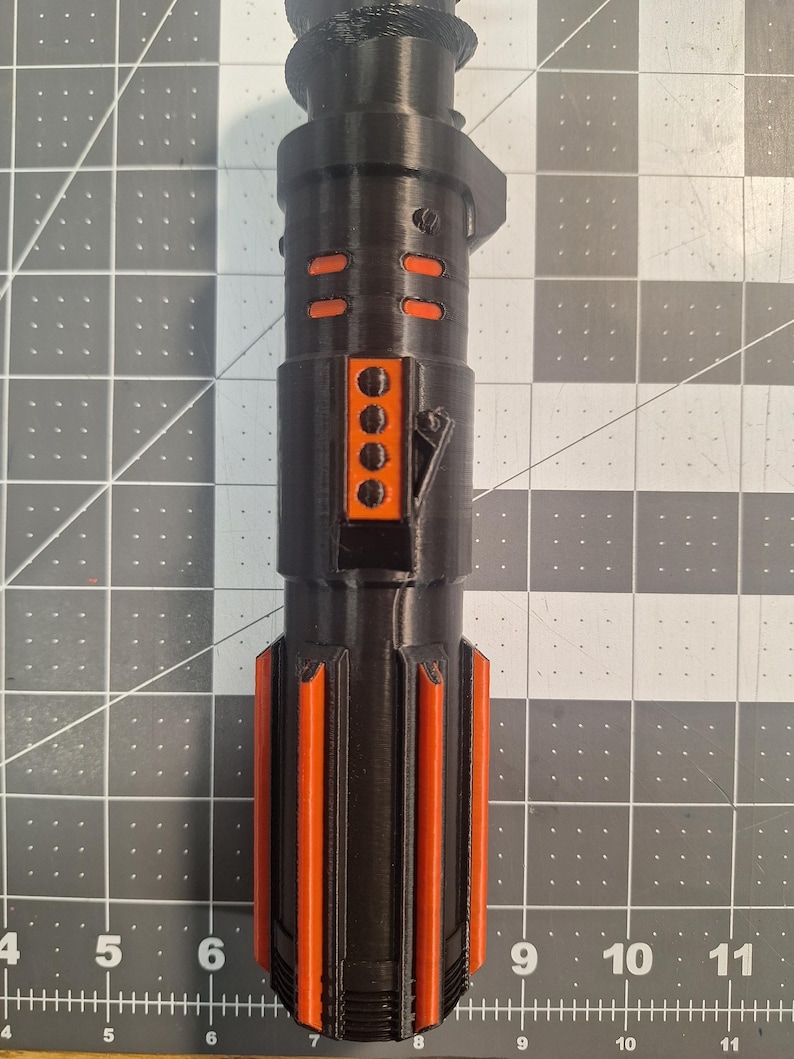May include: A black and orange lightsaber hilt, featuring a cylindrical design with orange accents. The hilt has a series of orange rectangular and vertical stripe details. The lightsaber is made of a 3D printed material.