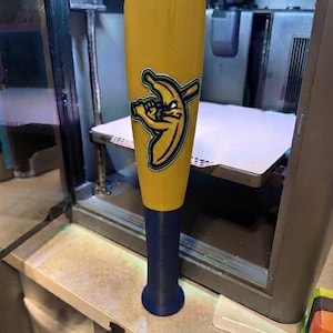 May include: A yellow baseball bat with a dark blue handle. The barrel features a graphic of a cartoon banana character holding a smaller baseball bat, outlined in dark green and black. The bat has a smooth, cylindrical shape with a flared knob.