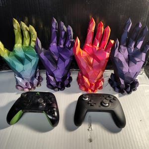 May include: Four geometric, crystal-like hand sculptures in gradient colors of yellow-green, purple, red-orange, and dark blue, displayed behind a black Xbox controller and a black Nintendo Switch Pro controller.