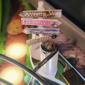May include: A whimsical signpost with four directional arrows. The arrows are labeled "Wrong Way," "Tea Party," "Turn Around," and "This Way." The signpost is white with a silver metal accent. The background includes a playing card and a blurred image.