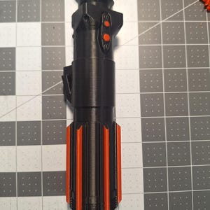 May include: A black and orange lightsaber hilt, featuring a cylindrical design with orange accents and two orange buttons. The hilt has a textured surface and is likely 3D-printed. The lightsaber is a prop or collectible item.