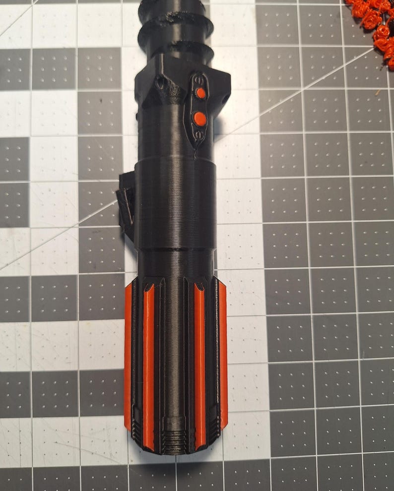 May include: A black and orange lightsaber hilt, featuring a cylindrical design with orange accents and two orange buttons. The hilt has a textured surface and is likely 3D-printed. The lightsaber is a prop or collectible item.