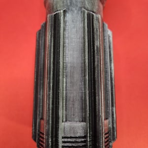 May include: A metallic gray, cylindrical object with vertical ribbed detailing and a flared base. The object has a textured surface and is set against a red background. The design suggests a mechanical or industrial aesthetic.