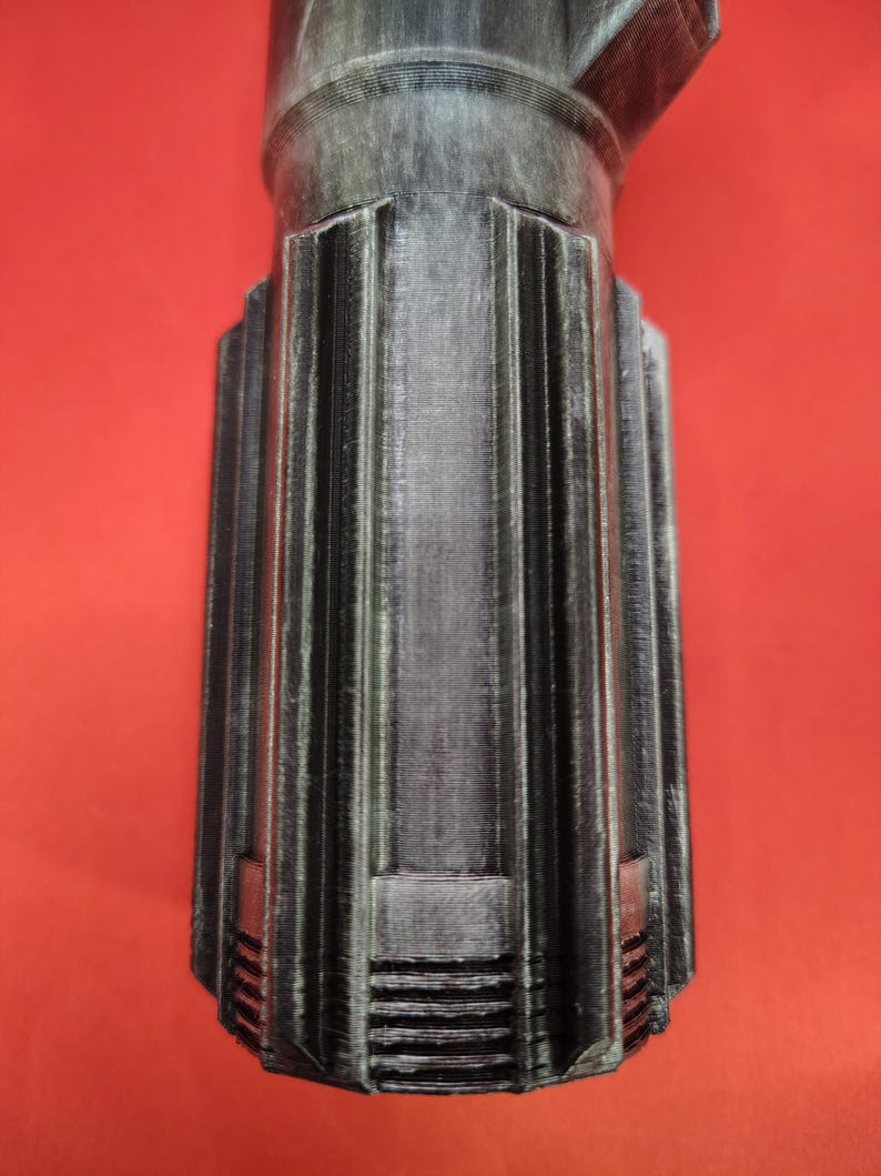 May include: A metallic gray, cylindrical object with vertical ribbed detailing and a flared base. The object has a textured surface and is set against a red background. The design suggests a mechanical or industrial aesthetic.