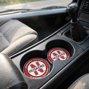 May include: Two round car coasters with the text "NEBRASKA FOOTBALL HUSKERS A WINNING TRADITION" in red and white. The coasters are in a car's cup holders, with a black gear shift and gray interior.