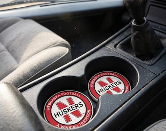 Peut inclure: Deux dessous de verre ronds pour voiture avec le texte "NEBRASKA FOOTBALL HUSKERS A WINNING TRADITION" en rouge et blanc. Les dessous de verre sont dans les porte-gobelets d'une voiture, avec un levier de vitesse noir et un intérieur gris.