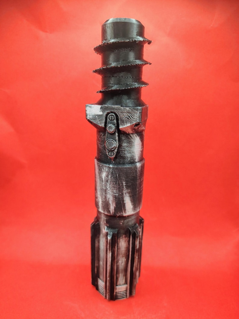 May include: A black and silver prop lightsaber hilt with a textured design. The hilt features a series of spiral ridges near the top, a detailed control section, and a segmented lower portion. The background is a solid red.