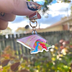 May include: A clear acrylic keychain featuring a colorful illustration of gummy worms spilling out of a pink bag. The bag has the text "Gummy Worms" and a rainbow. The keychain has a gold-colored clasp.