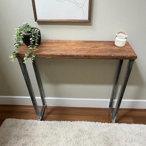 May include: A rustic console table with a stained wooden top and angled metal legs. The table is approximately 122 cm long and features a natural wood grain. A small white ceramic jar and a potted plant sit on the table.