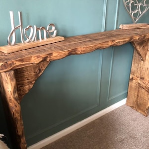 May include: Rustic wooden console table with a natural wood finish. The table has a single shelf and a wide, flat top. The table is decorated with a silver metal "Home" sign and a silver metal heart design.