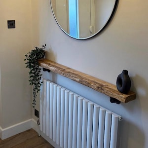 May include: A wooden shelf with a natural edge, supported by black brackets, is mounted above a white radiator. A black vase sits on the shelf, and a trailing green plant hangs down. A large round mirror hangs on the wall.
