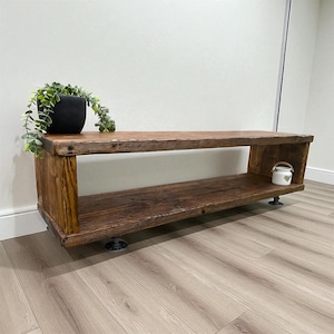 Puede incluir: Banco de madera r&uacute;stico con tinte oscuro. El banco tiene dos estantes y est&aacute; sostenido por patas de metal negro. Una maceta negra con una planta verde se encuentra en el estante superior, y una peque&ntilde;a jarra decorativa blanca est&aacute; en el estante inferior.