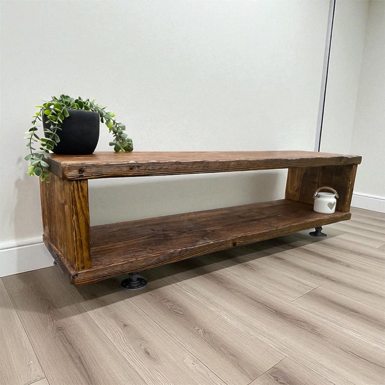 Puede incluir: Banco de madera r&uacute;stico con tinte oscuro. El banco tiene dos estantes y est&aacute; sostenido por patas de metal negro. Una maceta negra con una planta verde se encuentra en el estante superior, y una peque&ntilde;a jarra decorativa blanca est&aacute; en el estante inferior.