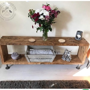 May include: Rustic wooden console table with two shelves and industrial pipe legs. The table is decorated with a bouquet of flowers, a decorative box, and a ceramic cat figurine.