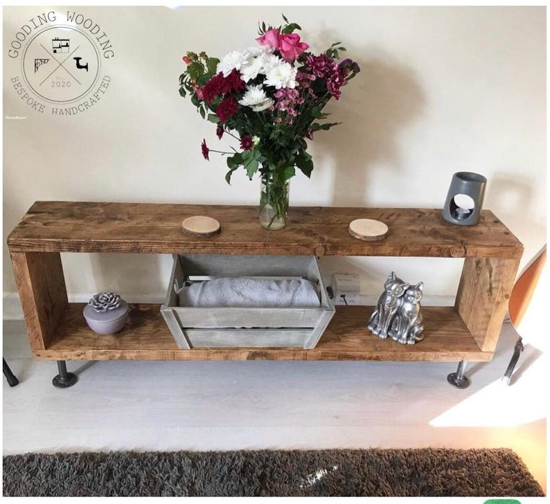 May include: Rustic wooden console table with two shelves and industrial pipe legs. The table is decorated with a bouquet of flowers, a decorative box, and a ceramic cat figurine.
