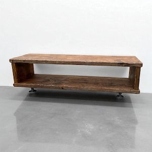 Puede incluir: Mesa de centro rectangular de madera r&uacute;stica con un estante inferior. La mesa es de madera marr&oacute;n oscuro y se apoya sobre cuatro peque&ntilde;os pies met&aacute;licos. El dise&ntilde;o presenta una construcci&oacute;n sencilla de estante abierto, adecuada para un sal&oacute;n. Mide unos 120 cm de largo.