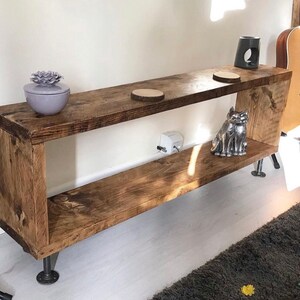 May include: A rustic wooden shelf unit with two shelves and metal pipe legs. The shelf is stained a dark brown color and has a natural wood grain. There are decorative items on the shelves, including a small ceramic planter with a purple flower, a wooden coaster, and a silver metal cat figurine.