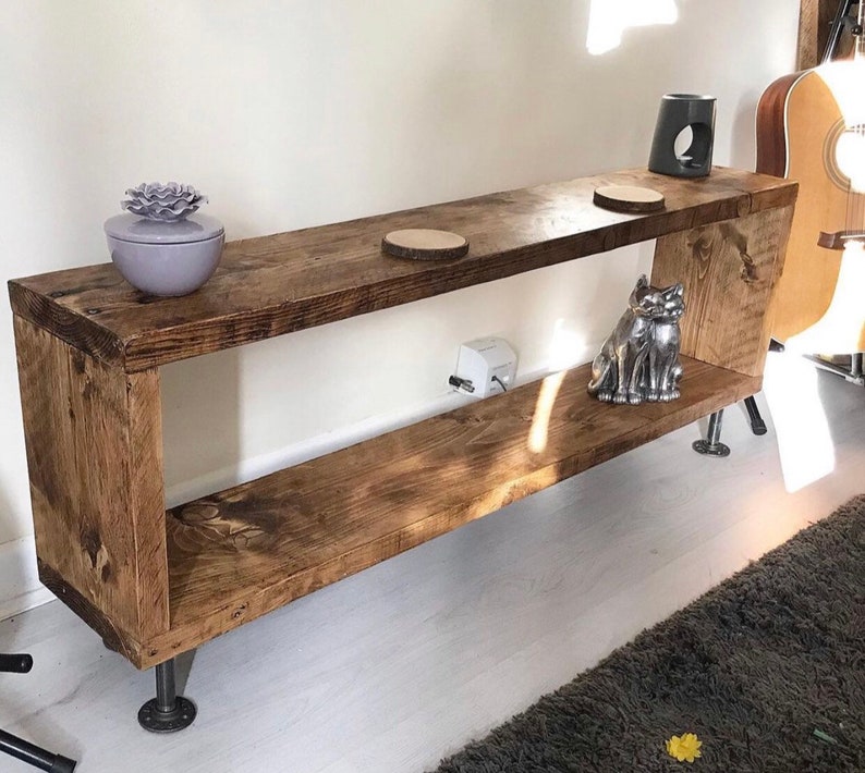 May include: A rustic wooden shelf unit with two shelves and metal pipe legs. The shelf is stained a dark brown color and has a natural wood grain. There are decorative items on the shelves, including a small ceramic planter with a purple flower, a wooden coaster, and a silver metal cat figurine.