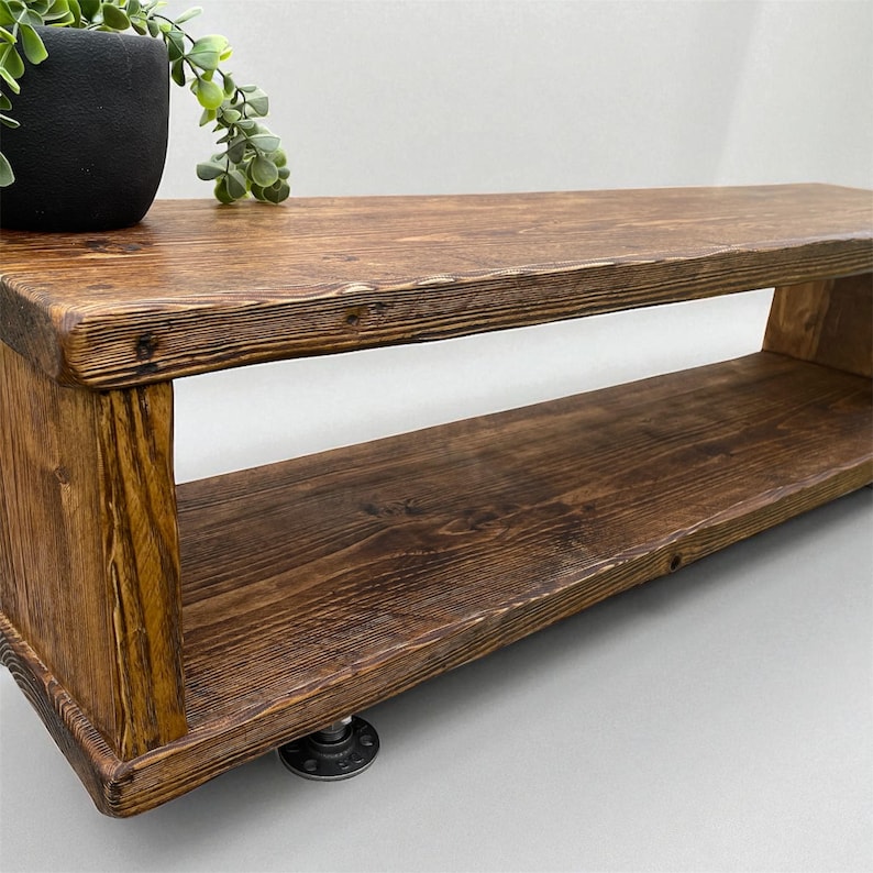 Puede incluir: Estanter&iacute;a rectangular de madera r&uacute;stica con tinte oscuro y dos niveles. Est&aacute; soportada por conexiones de tuber&iacute;as de metal negro. La veta de la madera es visible y los bordes est&aacute;n ligeramente redondeados. Una peque&ntilde;a maceta negra con vegetaci&oacute;n encima.