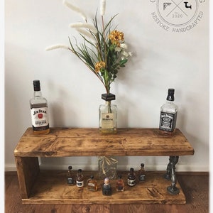 May include: Rustic wooden shelf with two tiers, featuring a pipe fitting design. The top tier has three bottles of liquor, while the bottom tier has a row of miniature liquor bottles. The shelf is stained in a dark brown color and has a distressed finish.