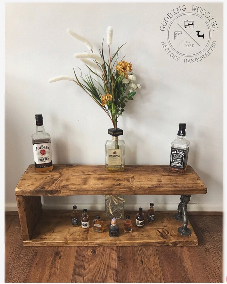 May include: Rustic wooden shelf with two tiers, featuring a pipe fitting design. The top tier has three bottles of liquor, while the bottom tier has a row of miniature liquor bottles. The shelf is stained in a dark brown color and has a distressed finish.