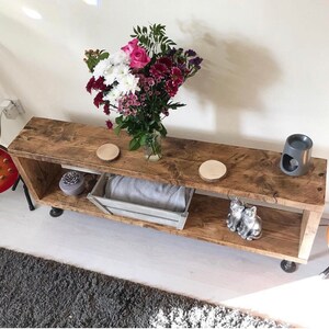 May include: A rustic wooden shelf unit with two shelves, featuring a bouquet of pink and purple flowers in a glass vase, two wooden coasters, a gray ceramic diffuser, and two silver cat figurines. The shelf is on black metal legs.