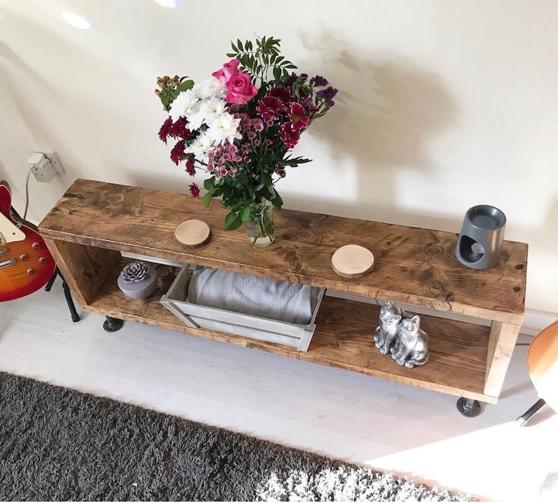 May include: A rustic wooden shelf unit with two shelves, featuring a bouquet of pink and purple flowers in a glass vase, two wooden coasters, a gray ceramic diffuser, and two silver cat figurines. The shelf is on black metal legs.