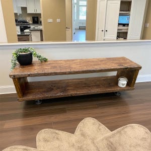 Puede incluir: Un banco de madera r&uacute;stico de dos niveles con un acabado marr&oacute;n oscuro. El banco tiene un estante inferior y est&aacute; soportado por ruedas de metal. Una peque&ntilde;a planta en maceta negra y un recipiente decorativo blanco se encuentran en el banco. El banco mide unos 150 cm de largo.
