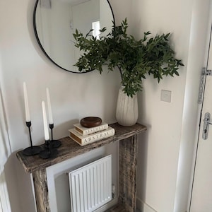 May include: A rustic wooden console table with a white radiator underneath. The table has a round mirror above it, a vase of green leafy plants, and two books stacked on top of each other with the words "Aesthetic" and "Abstract" on the covers.