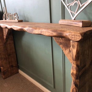 May include: A rustic wooden console table with a live edge top and two chunky legs. The table is stained a dark brown colour and has a natural wood finish. The table is decorated with a silver heart-shaped sculpture and a wooden sign that says "home".