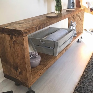 May include: A rustic wooden shelf unit with two shelves and metal pipe legs. The shelf is stained a dark brown and has a distressed finish. A gray wooden crate with a blanket inside sits on the lower shelf. A small decorative item sits on the upper shelf.