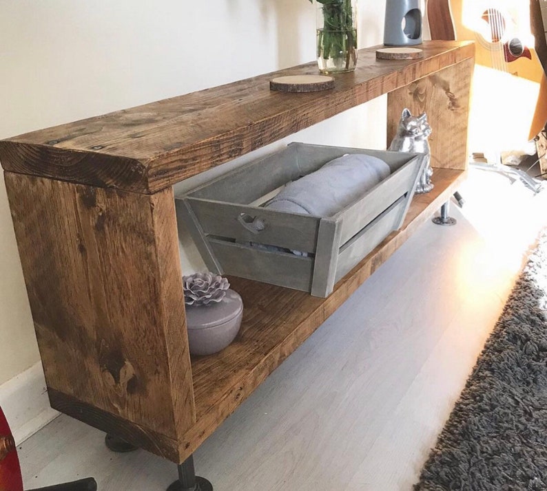 May include: A rustic wooden shelf unit with two shelves and metal pipe legs. The shelf is stained a dark brown and has a distressed finish. A gray wooden crate with a blanket inside sits on the lower shelf. A small decorative item sits on the upper shelf.