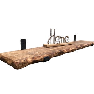 May include: A rustic wooden shelf with a live edge and black metal brackets. The shelf is decorated with a silver metal sign that says "Home".
