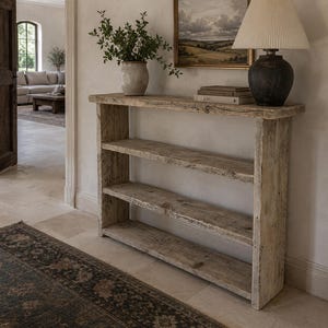 May include: A tall, narrow console table crafted from light-toned, weathered wood with a visible grain. The piece features three horizontal shelves, including the top surface, supported by two vertical side panels. It has a rectangular, open-frame design.